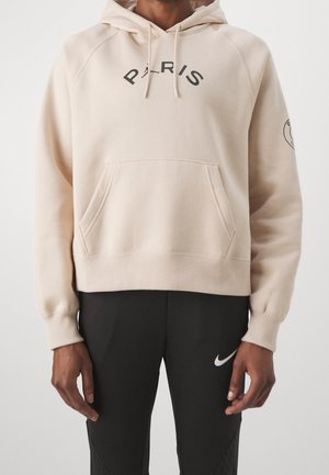 Personne portant un sweat à capuche beige avec le texte "PARIS" et un pantalon Nike noir avec un logo swoosh blanc, debout devant un fond uni.