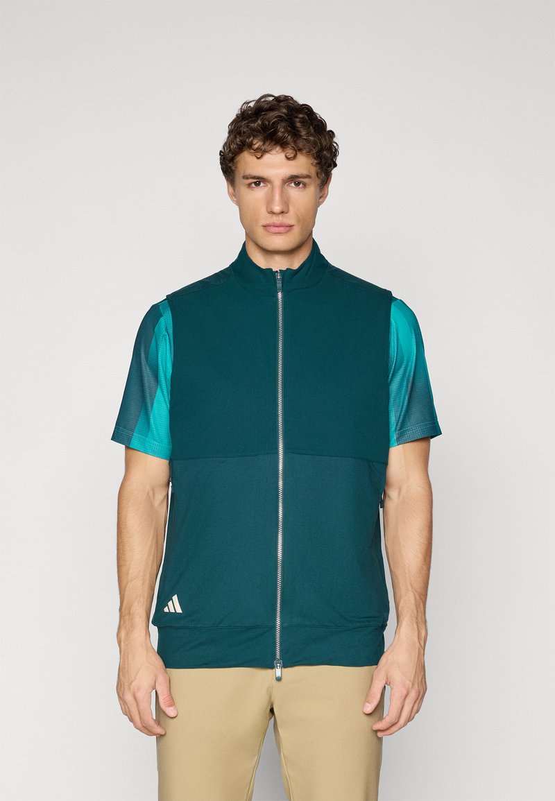 Young man wearing a teal zip-up sleeveless vest over a turquoise short-sleeve shirt and beige pants, standing against a plain background.