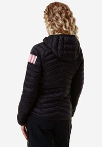 Giubbotto nero invernale con cappuccio, caratterizzato da cuciture diagonali e un patch della bandiera americana sulla manica sinistra. Tessuto lucido e liscio.