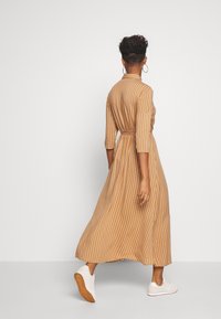 Mujer caminando de espaldas con un vestido midi de rayas verticales en tonos beige y blanco, de mangas tres cuartos y zapatillas blancas, sobre un fondo liso.