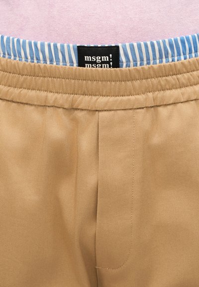 Μπεζ παντελόνι με ελαστική μέση πάνω από επένδυση με μπλε και λευκές ρίγες, που εμφανίζει μια μαύρη ετικέτα με το λογότυπο "msgm!" σε λευκό κείμενο.
