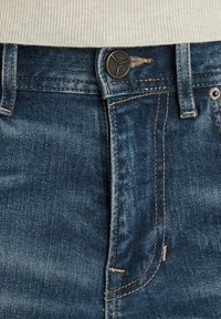 Vaqueros de mezclilla en azul oscuro con una textura desgastada, presentan un botón de metal, costuras naranjas en contraste y un diseño tradicional de cinco bolsillos.