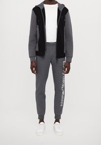 Czarna i szara bluza z zamkiem na dłogim rękawie, na białej koszulce, w zestawie z szarymi dresami z białym nadrukiem logo, uzupełnione białymi trampkami.