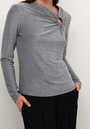 Femme portant un haut métallique argenté à manches longues avec un détail de découpe noué près du col et un pantalon noir, mains dans les poches.