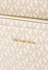 Fehér, texturált anyagú táska, amelyen egymásba kapcsolódó háromszögek mintája látható, arany cipzárral és arany leheletű logóval, amelyen a "Michael Kors" szöveg olvasható.