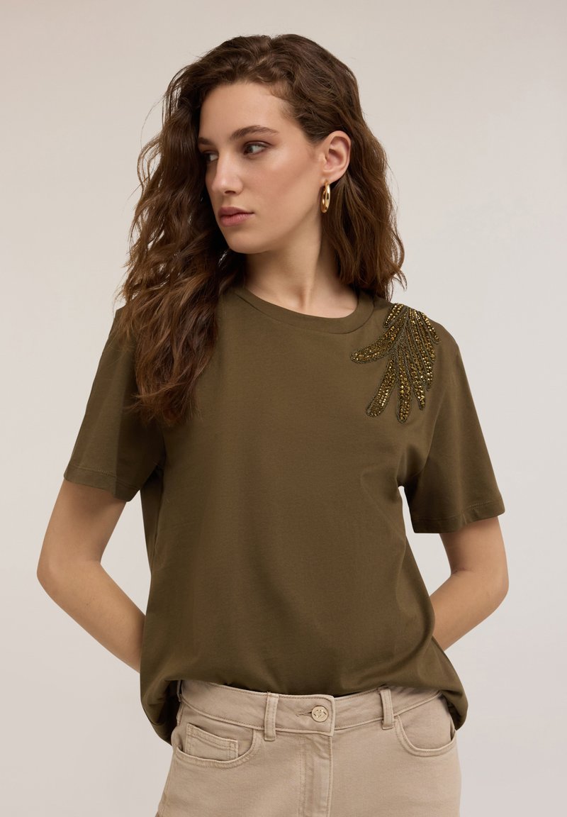 Camiseta de algodón verde oliva con un diseño de hoja de lentejuelas en el hombro izquierdo, de manga corta y corte holgado.