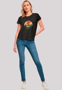 Black short-sleeve t-shirt featuring a circular graphic of mountains and trees in orange and green hues. Paired with blue jeans.