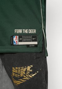 Maglietta da basket verde con il testo "FEAR THE DEER" e tag NBA, abbinata a pantaloni della tuta grigi con un logo Nike dorato.