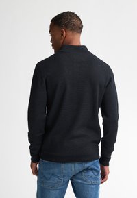 Maglione nero a maglia con colletto, caratterizzato da un tessuto texturizzato. Indossato con jeans in denim blu, mostrando la vista da dietro.