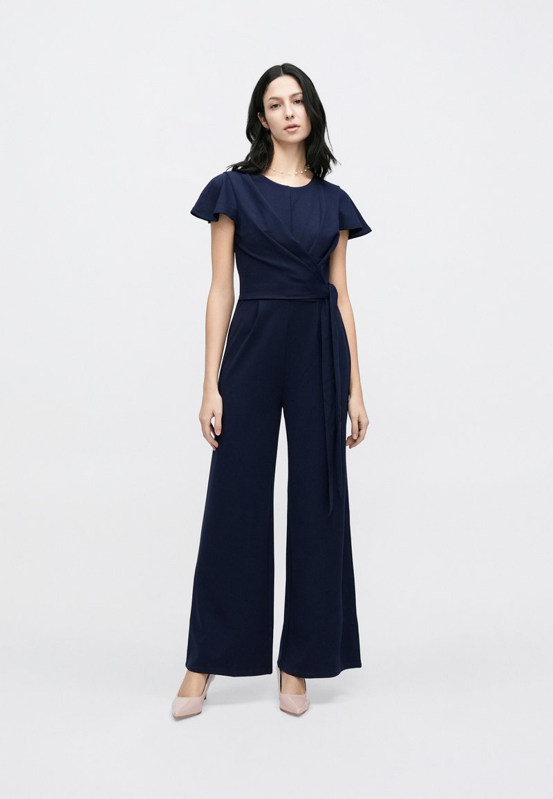 Mono azul marino con mangas cortas y volanté, delantero cruzado y cintura ajustable con lazo. El diseño de pierna ancha añade fluidez y comodidad.