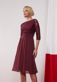 Robe bordeaux avec un corsage en dentelle et des manches trois-quarts, jupe en chiffon fluide et détail de taille froncée. Fond rouge et blanc visible.