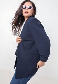 Marineblauwe blazer met een getailleerde pasvorm, voorzien van lange mouwen, een enkele borstzak en een soepele stof. Draag het met ronde, roze zonnebril.