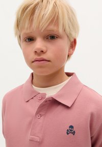 Jasnoróżowa koszulka polo z teksturowanej tkaniny, z ciemnoniebieskim logo czaszki i skrzyżowanych kości na lewej piersi oraz z zapięciem na dwa guziki.