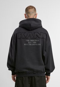 Fekete kapucnis pulóver texturált "KANU" logóval, alapítási évvel és védjegy szöveggel a hátsó oldalon. Bordás mandzsetta és szegély. Puha anyag.