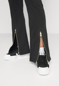 Čierne tepláky so zlatými zipsami na členkoch, skombinované s čierno-bielymi teniskami s textúrovaným povrchom a gumovou podrážkou.