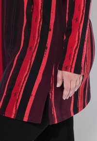 Long-sleeve tunic featuring red and black stripes with a side slit and rounded hem. Made of soft material, it offers a relaxed fit.