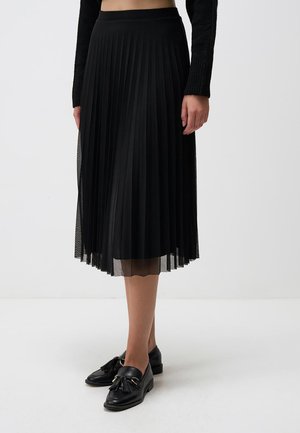 Frau trägt einen schwarzen plissierten Midirock, ein schwarzes langärmeliges Oberteil und schwarze Loafers mit Quasten, steht vor einem einfarbigen weißen Hintergrund.