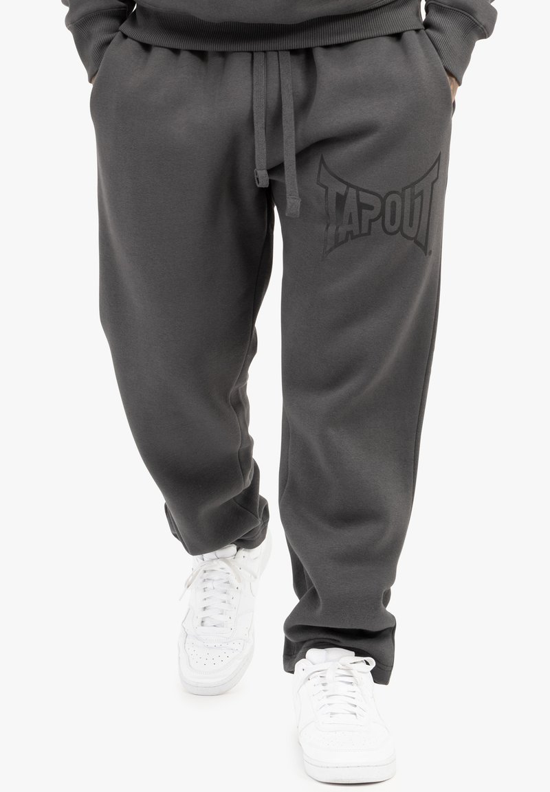 Grå joggingbukser i bomuldsblanding med snoretræk i taljen og et stort, sort TAPOUT-logo på venstre lår, parret med hvide sneakers.