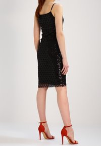Černé krajkové šaty s přiléhavým střihem a diamantovými vzory. Kombinované s červenými lodičkami na vysokém podpatku a se sponou kolem kotníku. Zobrazen pohled zezadu.