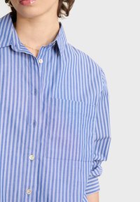 Blue and white striped button-up shirt with a collar, featuring a single chest pocket and rolled sleeves. Smooth fabric texture.