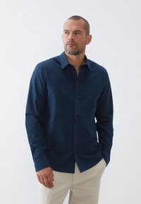 Camisa de botones azul marino con un sutil patrón de puntos. Presenta un cuello estándar y mangas largas, combinado con pantalones de color claro.