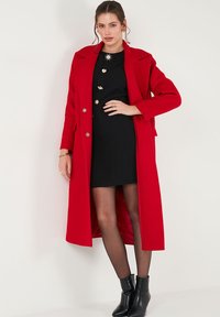 Long red wool coat with a structured design, notched lapels, and double-breasted buttons, worn over a black ribbed dress and black ankle boots.