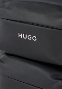 Sort rygsæk lavet af tekstureret stof, med lynlåslukning og et fremtrædende sølvfarvet "HUGO" logo på fronten.