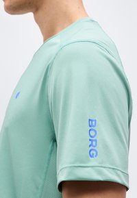 Κοντινό πλάνο ενός ατόμου που φοράει ανοιχτοπράσινο αθλητικό κοντομάνικο πουκάμισο με την επιγραφή "BORG" σε μπλε χρώμα στο μανίκι.