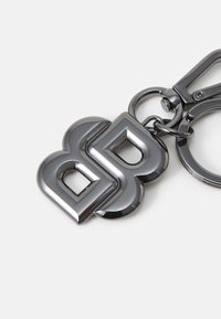 Portachiavi in metallo di un elegante grigio metallizzato lucido, caratterizzato da un logo stilizzato con la doppia "B" e un robusto gancio per il fissaggio. Texture liscia, design elegante.