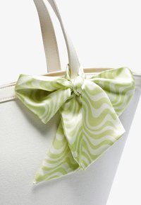 Hvid tote bag lavet af tekstureret materiale, med en lysegrøn satinbue med bølgede mønstre, der er fastgjort øverst.