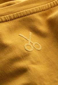 Geel stof met een gestructureerd oppervlak, voorzien van een geborduurd schaarontwerp in een lichtere tint, subtiel geïntegreerd in het materiaal.