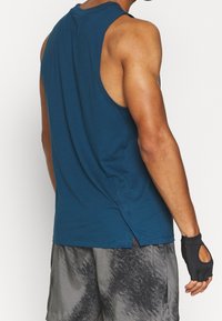Blauwe mouwloze sporttop met een ontspannen pasvorm, voorzien van een ronde halslijn en zijsplitten, gecombineerd met grijze gebroken shorts en vingerloze handschoenen.