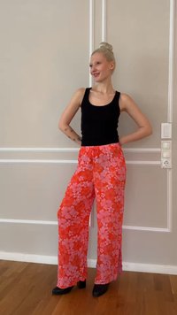 Γυναίκα με ξανθό κότσο που φοράει μαύρο τοπ χωρίς μανίκια, ζωηρό κόκκινο φλοράλ παντελόνι με φαρδιά πόδια και μαύρα παπούτσια, που στέκεται σε εσωτερικό χώρο μπροστά σε μπεζ τοίχο.