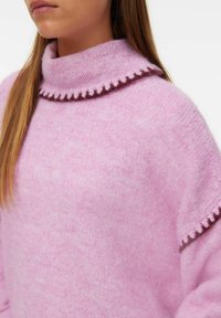 Pull en tricot rose avec col montant, présentant une bordure brune à volants et un tissu doux et texturé.
