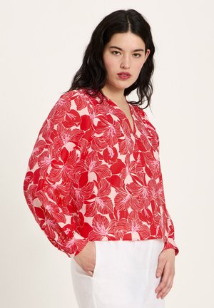 Junge Frau mit langen dunklen Haaren, die eine rot-rosa geblümte Langarmbluse und weiße Hose trägt, mit einer Hand in der Tasche stehend.