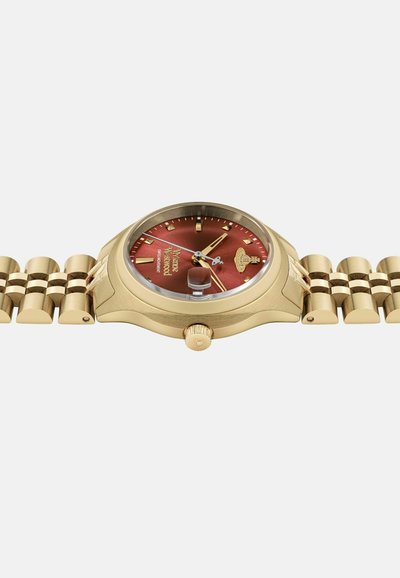 Reloj de pulsera en tono dorado con un dial rojo redondo, índices y manecillas doradas. Presenta una pulsera metálica pulida y una corona visible en el costado.