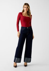 Rode top met lange mouwen en vierkante halslijn, gecombineerd met hooggetailleerde, wijd uitlopende donkere spijkerbroek met contrasterende spijkerboorden. Bruine hakken.