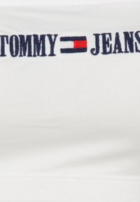 Biała prążkowana tkanina z granatowym haftowanym napisem "TOMMY JEANS"; posiada czerwony i niebieski prostokątny akcent pomiędzy słowami.