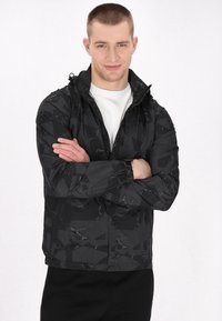 Jachetă neagră cu fermoar, cu un model geometric, având mansete elastice și glugă. Fabricată dintr-un material ușor, rezistent la apă.