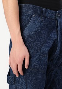 Mano descansando al lado de pantalones de mezclilla azul oscuro con un patrón texturizado abstracto y un bolsillo delantero, combinados con una camiseta negra.