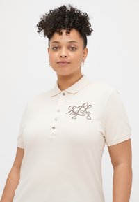 Camisola polo em tecido bege claro com colarinho, mangas curtas e um logótipo decorativo bordado em fio escuro no peito. Cinco botões.