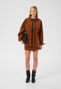 Veste chemise en velours côtelé marron avec deux poches poitrine, associée à une mini-jupe assortie. Le mannequin tient un sac vert foncé et porte des bottines noires.