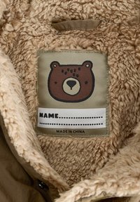 Brun fluffig inredning med en mjuk textur. Beige etikett med ett tecknat björnhuvud och ett tomt namnfält, med texten "MADE IN CHINA".