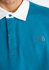 Zöldeskék póló fehér gallérral, kék gombbal és a The North Face hímezett logójával a bal mellen. Puha, textúrált anyag.