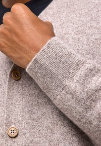 Jasnobrązowy kardigan wykonany z miękkiego, teksturowanego materiału, z prążkowanymi mankietami i drewnianymi guzikami. Zbliżenie na detal rękawa i pozycję dłoni.