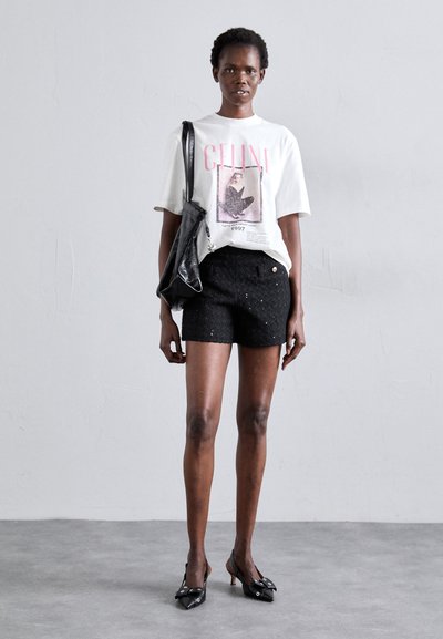 T-shirt blanc avec un imprimé graphique et un texte rose, shorts en tweed noir avec un bouton à l'avant, sac à main noir et escarpins à bout pointu.
