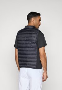 Μαύρο γεμιστό γιλέκο με ψηλό γιακά, με ραφές και κομψό σχέδιο, φορεμένο πάνω από ένα μαύρο κοντομάνικο πουκάμισο.