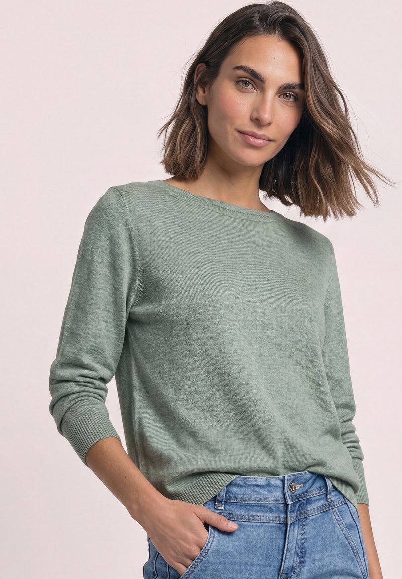 Femme aux cheveux châtains mi-longs, portant un pull vert clair et un jean bleu, debout avec une main dans la poche, sur un fond uni.