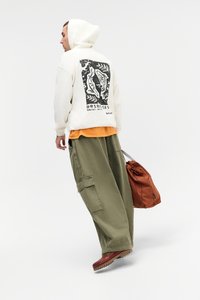 Witte hoodie met een grafische print op de achterkant, gedragen over een oranje shirt. Olijfgroene cargobroek en een oranje geruit rugzak met trekkoord.