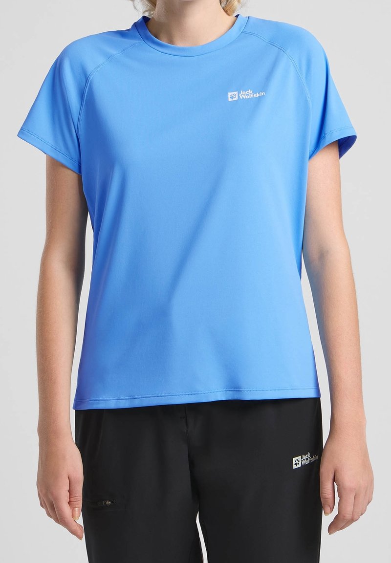 Persona vistiendo una camiseta deportiva de manga corta azul claro y pantalones negros, ambos con logotipos de Jack Wolfskin en el pecho y el muslo.